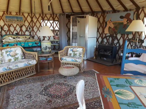 Cozy yurt interior with wicker furniture, a bed, a small fridge, and a decorative rug. Warm, inviting atmosphere.