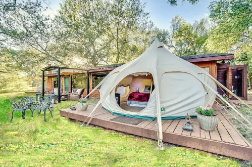 A cozy glamping tent on a wooden deck surrounded by greenery and a rustic cabin in the background.