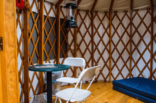 Interior of a yurt featuring a table, two chairs, a lantern, and a mattress on a wooden floor.