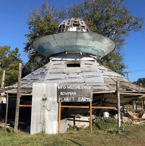 A weathered UFO-shaped structure with a sign reading "UFO Welcome Center" in a rural setting. Trees in the background.