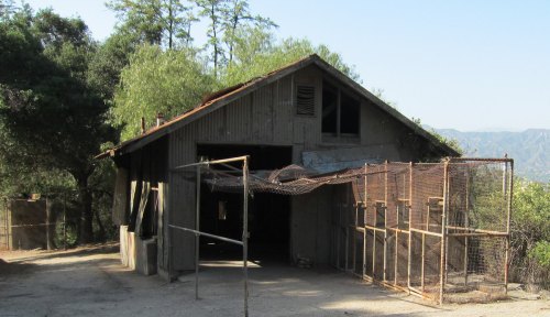 A weathered barn with a fenced area, surrounded by trees and mountains in the background under a clear blue sky.
