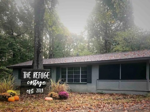 A cozy cottage surrounded by trees, featuring a sign that reads "The Refuge Cottage est. 2019" and autumn decorations.