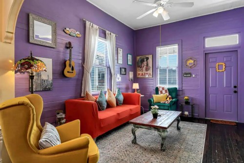 Colorful living room with purple walls, a red sofa, yellow armchair, and eclectic decor, featuring a guitar and vintage lamp.