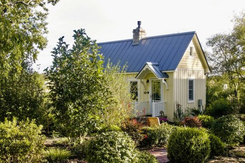 A charming yellow cottage surrounded by lush greenery and colorful flowers under a clear sky.