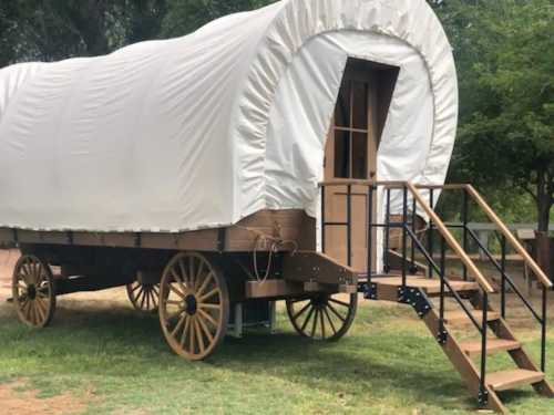 A covered wagon with wooden wheels and a staircase, set on grassy ground under a tree.