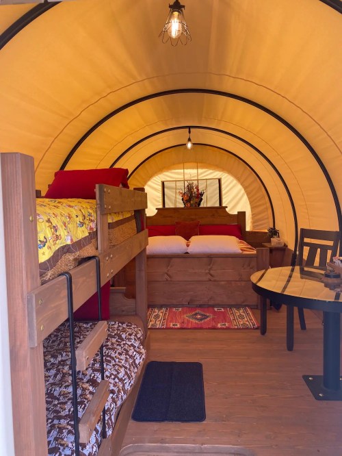 Cozy interior of a glamping tent featuring a bunk bed, a double bed, and a small dining table. Warm lighting enhances the ambiance.