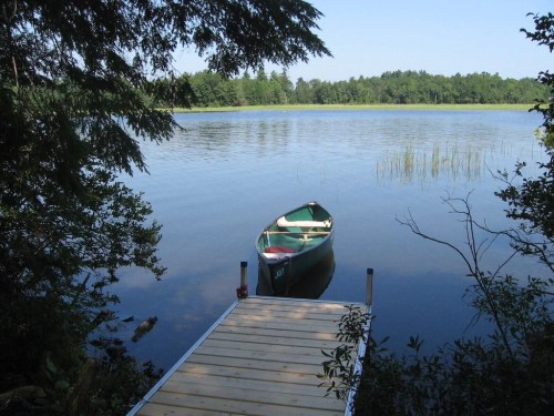 A calm lake scene with a wooden dock and a green canoe moored nearby, surrounded by trees and clear blue skies.