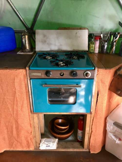 A vintage blue gas stove with an oven, set in a rustic kitchen with wooden surfaces and various kitchen utensils nearby.