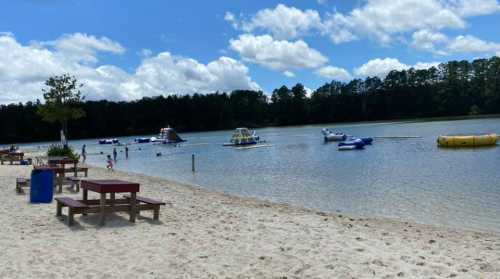 A sandy beach by a lake with inflatable water toys and people enjoying the water under a blue sky with clouds.