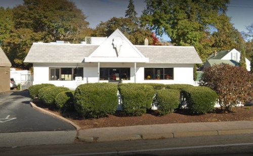 A white building with a peaked roof, surrounded by neatly trimmed bushes and trees, located on a street.