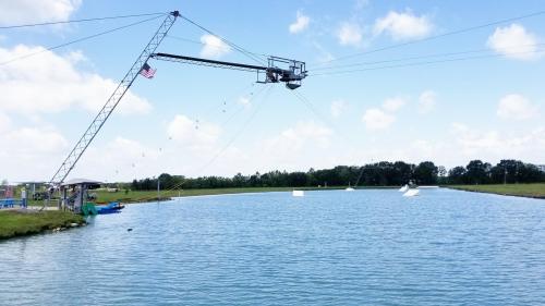 A cable park with a crane over a calm lake, featuring a blue sky and scattered clouds.