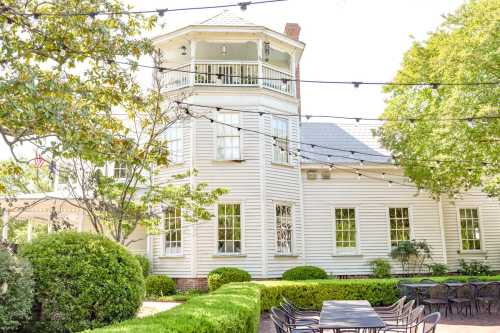 A charming white house with a tower, surrounded by greenery and outdoor seating under string lights.