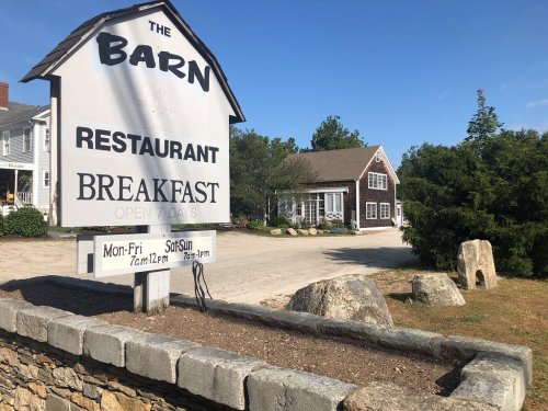 Sign for "The Barn" restaurant, featuring breakfast hours, with a rustic building and trees in the background.