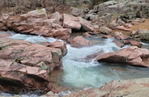 A flowing river cascades over large, smooth pink and gray rocks in a natural landscape.