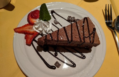 A slice of chocolate mousse cake topped with chocolate drizzle, whipped cream, strawberries, and a mint leaf on a white plate.