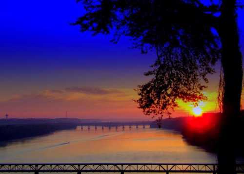 Sunset over a river, with vibrant colors in the sky and a silhouette of trees and a bridge in the foreground.