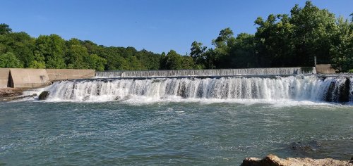 A serene waterfall cascades over a dam, surrounded by lush green trees under a clear blue sky.