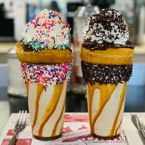 Two large milkshakes topped with whipped cream, sprinkles, and chocolate, served in donut-shaped cups.