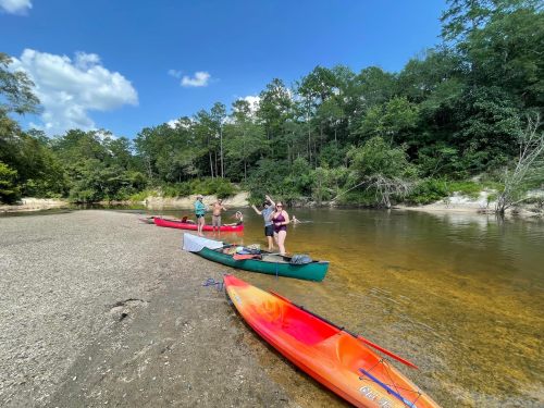 Three people on a sandy riverbank with kayaks, surrounded by trees and a clear blue sky.