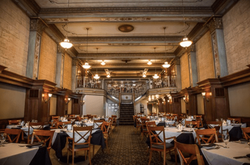 Elegant restaurant interior with wooden furniture, high ceilings, and decorative lighting, featuring tables set for dining.