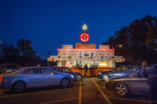 A brightly lit food stand at dusk, surrounded by parked cars, with a menu displayed prominently on the front.