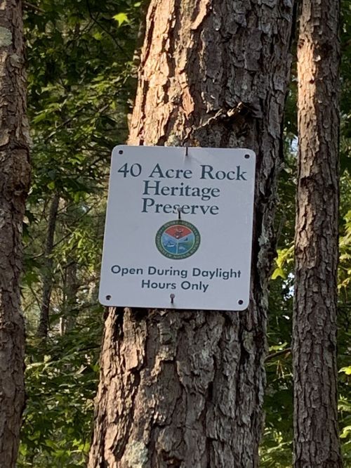 Sign on a tree reading "40 Acre Rock Heritage Preserve - Open During Daylight Hours Only." Surrounded by greenery.