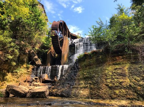A watermill with a large wooden wheel, surrounded by lush greenery and cascading water over rocky terrain.