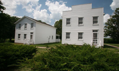 Two white, wooden houses with green shrubs in the foreground, set against a partly cloudy sky.