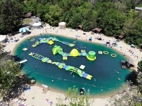 Aerial view of a sandy beach with inflatable water attractions and people enjoying a sunny day at a lake.
