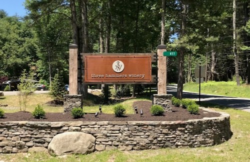 Sign for Three Hammers Winery, featuring a wooden design, surrounded by greenery and a stone wall.