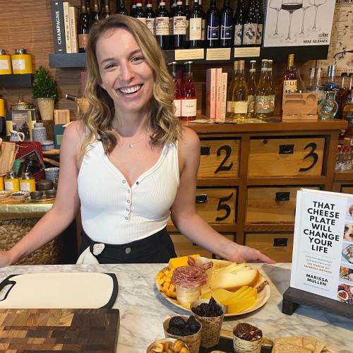 A smiling woman stands in a kitchen with a cheese platter, wine bottles, and a cookbook titled "That Cheese Plate Will Change Your Life."