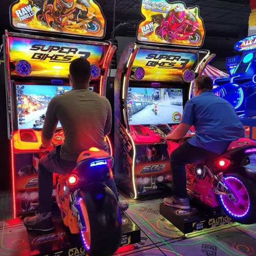 Two people playing motorcycle racing arcade games, illuminated by colorful lights in a gaming arcade.
