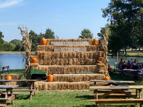 A rustic display of hay bales and pumpkins at Cedar Lake Cellars Winery, near a serene lake and picnic tables.