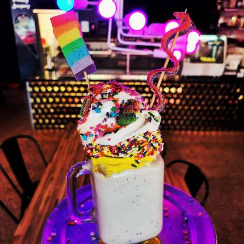 A colorful milkshake topped with whipped cream, sprinkles, and a rainbow flag, served in a glass mug.