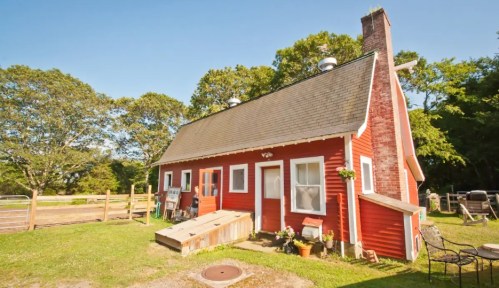 A charming red farmhouse with a sloped roof, surrounded by greenery and a wooden fence on a sunny day.