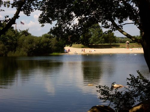 A serene lake view framed by trees, with a sandy beach and people relaxing in the background under a clear blue sky.