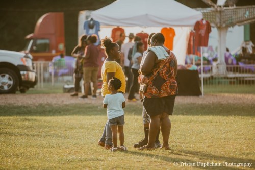 A woman and two children converse outdoors at a lively event, with tents and people in the background.