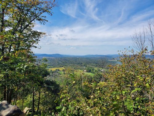A scenic view of rolling hills and valleys under a blue sky with scattered clouds, framed by trees in the foreground.