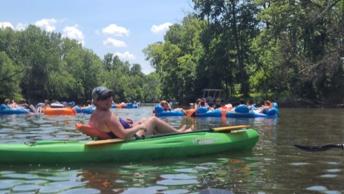 A man in a kayak enjoys a sunny day on a river, surrounded by people floating on colorful inner tubes.