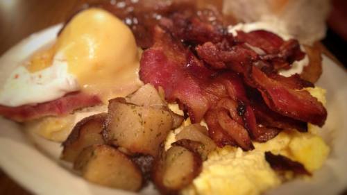 A plate of breakfast featuring eggs Benedict, crispy bacon, scrambled eggs, and seasoned potatoes.