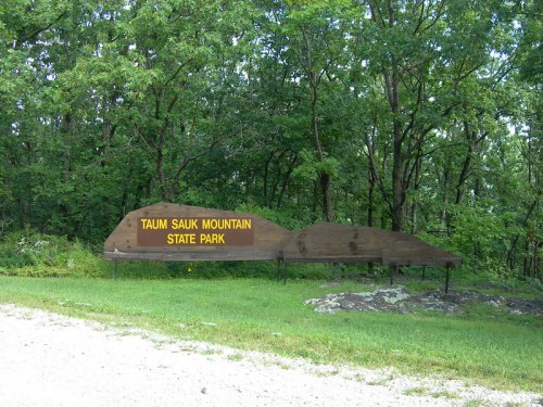 Sign for Taum Sauk Mountain State Park, surrounded by greenery and rocky terrain.