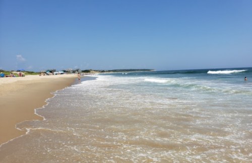 A serene beach scene with gentle waves, golden sand, and a clear blue sky. People enjoy the shoreline in the distance.