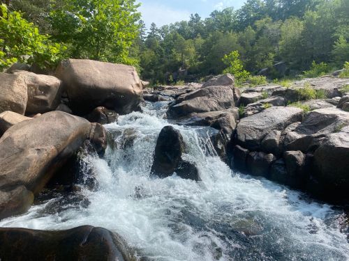 A flowing river cascades over large rocks, surrounded by lush green trees and a clear blue sky.