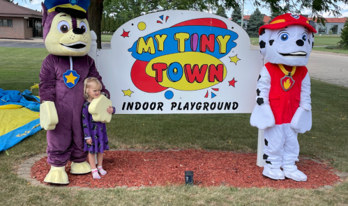 A young girl poses with two costumed characters in front of a colorful sign for "My Tiny Town Indoor Playground."
