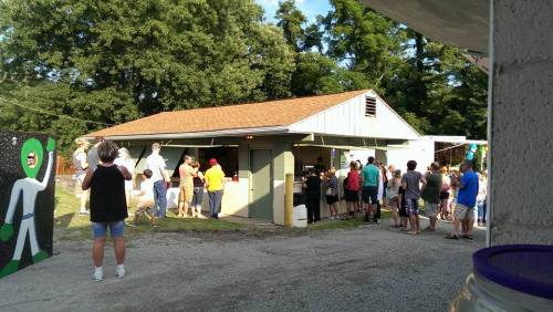 A crowd gathers outside a small food stand with a green alien decoration, surrounded by trees on a sunny day.