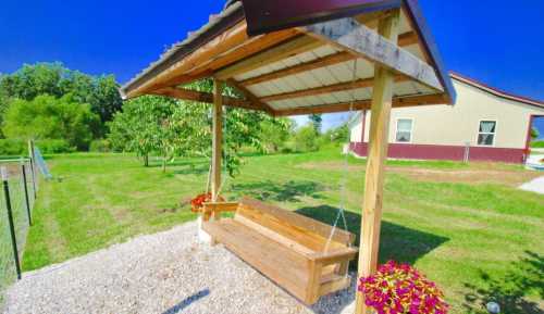 A wooden swing under a shelter, surrounded by green grass and trees, with a house in the background.