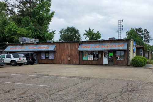 A rustic restaurant with a wooden exterior, featuring signs for food and drinks, surrounded by trees and parked cars.