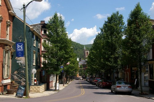 A tree-lined street in a small town, featuring historic buildings and parked cars under a blue sky with fluffy clouds.