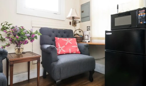 Cozy corner with a gray armchair, a small table, a microwave, and a mini fridge, featuring a vase of flowers.