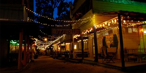 A charming street lit with string lights at night, featuring wooden buildings and a festive atmosphere.